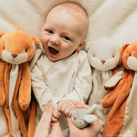 Baby laughs, models Little Sunshine Romper and is surrounded by plush bunnies
