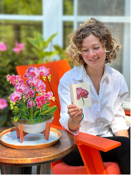 Woman sits in chair with Orchid Oasis greeting card on table next to her as she reads her card