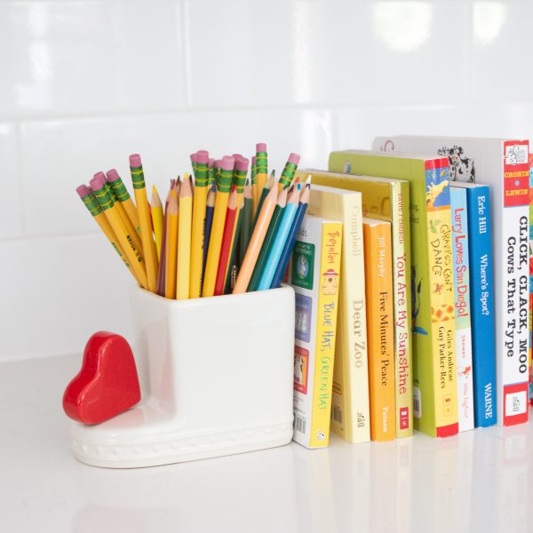 #A023 - Be Mine - heart mini by Nora Fleming posed on Cutie Container filled with pencils, propping up books